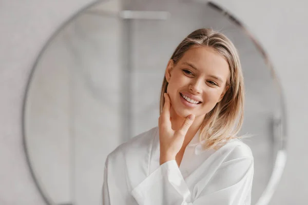 Mujer bastante caucásica mirando al espejo, tocando su piel y sonriendo a su reflejo, de pie en el baño — Foto de Stock