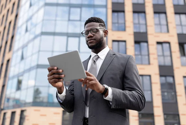 Фокусований чорний чоловік у формальному носінні, що стоїть біля офісної будівлі, тримає цифровий планшет, використовуючи бізнес-додаток — стокове фото