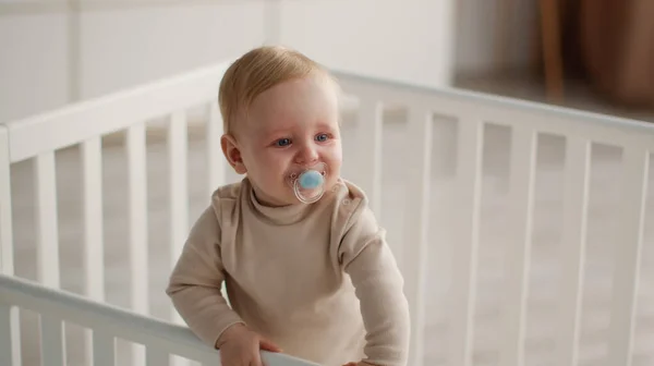 Lugnande för småbarn. Liten bebis med napphållare stående i spjälsäng, tittar åt sidan på sovrum, panorama, ledigt utrymme — Stockfoto
