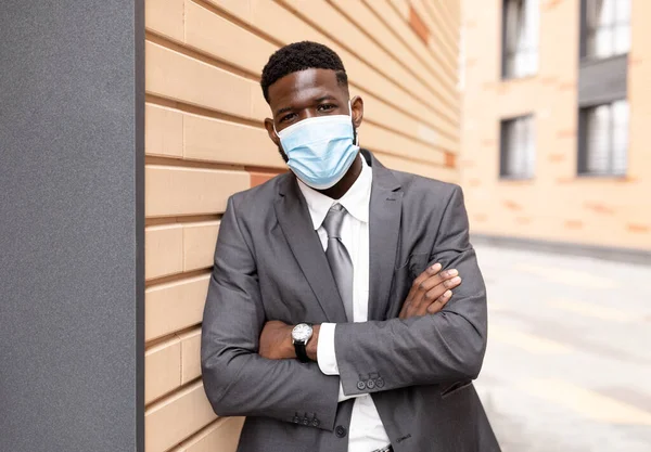 Retrato de empreendedor afro-americano em máscara protetora médica posando ao ar livre perto do centro de negócios, espaço livre — Fotografia de Stock