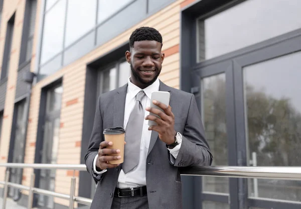 Χαμογελώντας Αφροαμερικανός επιχειρηματίας σε επίσημη ένδυση με καφέ για να πάει και smartphone στέκεται δίπλα στο κτίριο γραφείων — Φωτογραφία Αρχείου