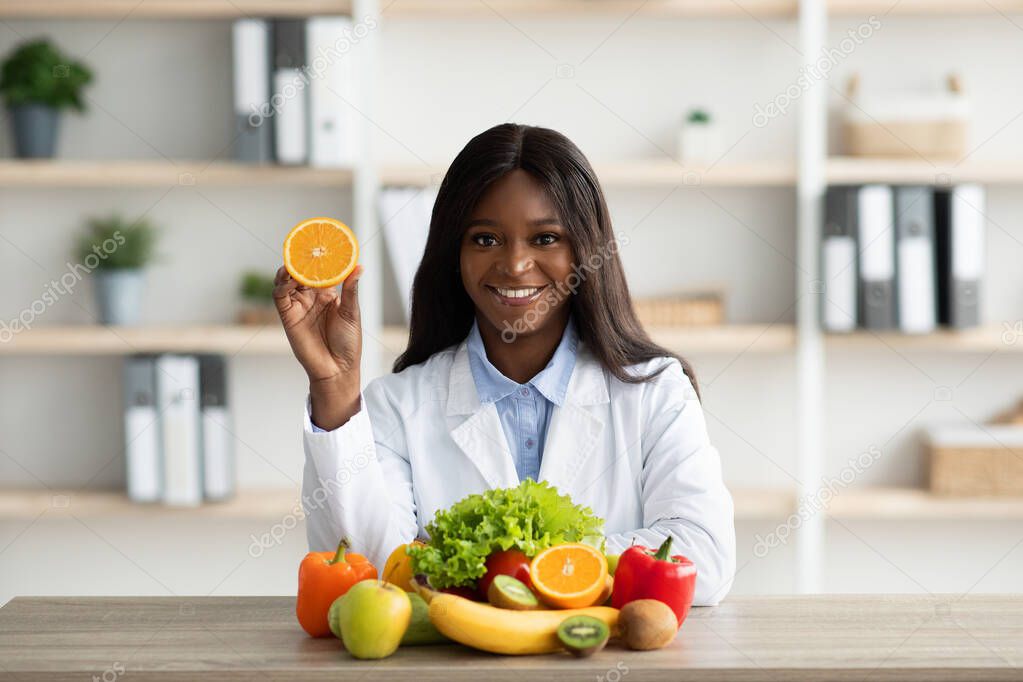 Start your day with fresh fruits. Happy black lady nutritionist holding orange half, sitting in office and smiling