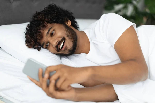 Sonriente chico hindú acostado en la cama con teléfono inteligente — Foto de Stock