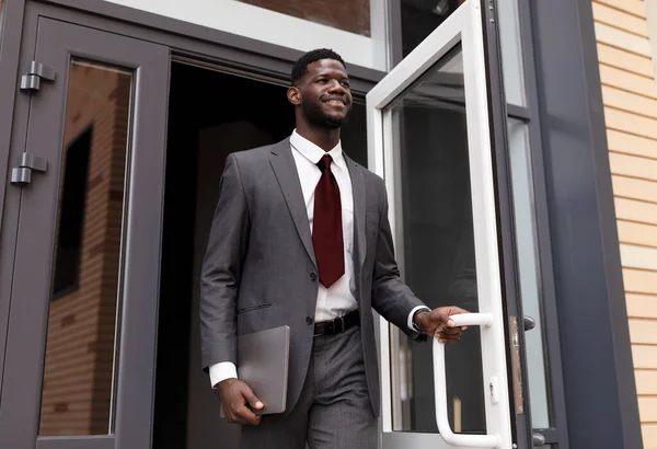 Självsäker afrikansk amerikansk affärsman i formell kostym går ut från moderna kontor centrum och ler — Stockfoto