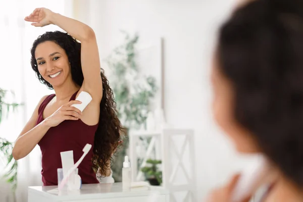 Wanita muda cantik menerapkan deodoran, berdiri di depan cermin di rumah, menyalin ruang. Konsep kebersihan pribadi — Stok Foto