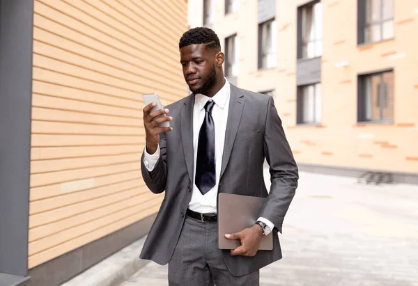 Young black CEO checking emails on cellphone, walking outdoors with laptop in hand, having break outside