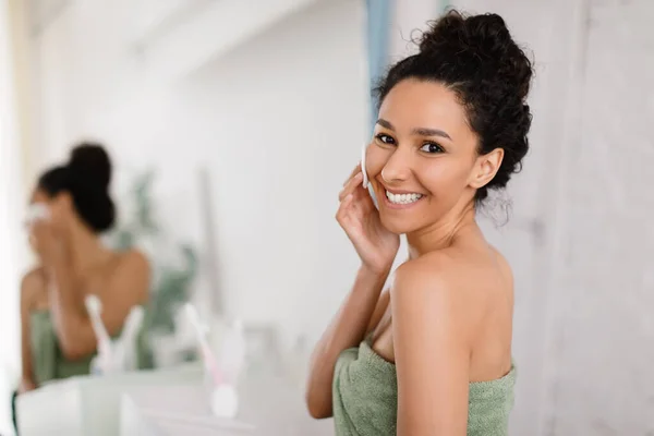 Perfekte Haut. Fröhliche junge brünette Frau in Handtuch berührt Gesicht nach Bad oder Dusche, in der Nähe Spiegel im Badezimmer — Stockfoto