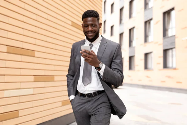 Africký generální ředitel kráčí vedle kancelářské budovy, používá smartphone a usmívá se, používá aplikaci nebo chatuje s klienty — Stock fotografie