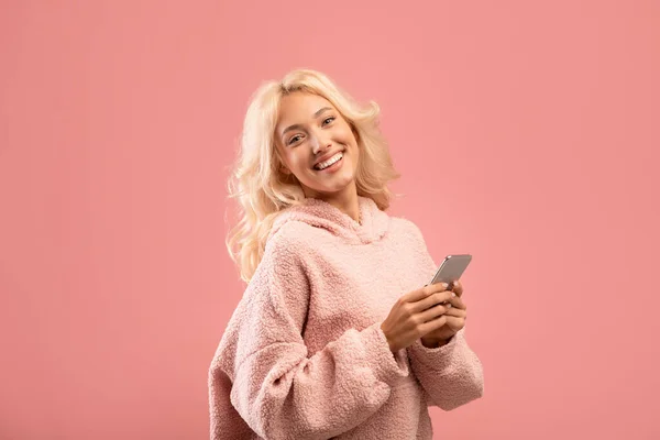 Люди і технології концепції. Досить молода леді, використовуючи смартфон і посміхаючись на камеру, рожевий студійний фон — стокове фото