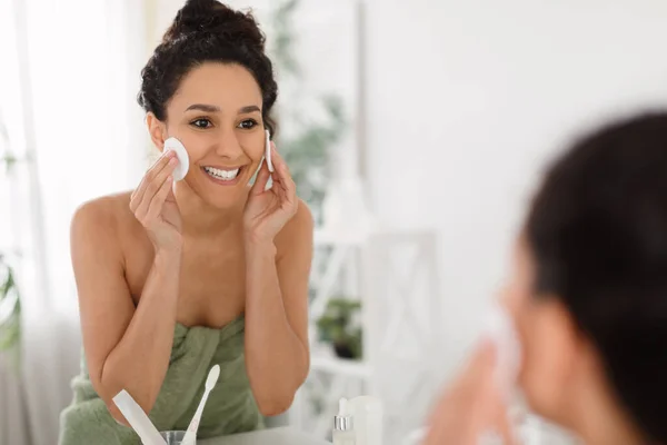Positive woman removing makeup with cotton pads, applying skin tonic, looking at mirror, enjoying her facial products