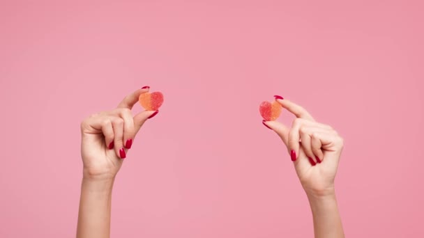 Manos femeninas sosteniendo dos caramelos de gelatina en forma de corazón, fondo rosa, recortado — Vídeos de Stock