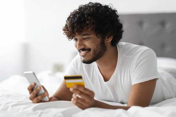 Tmavý chlap ležící v posteli s kreditní kartou a smartphonem — Stock fotografie