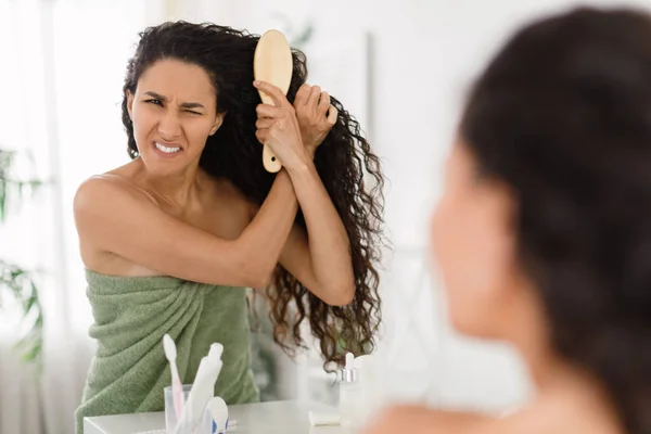 Frustrated young lady having problem brushing her tangled hair in front of mirror at home, copy space — Stock Photo, Image