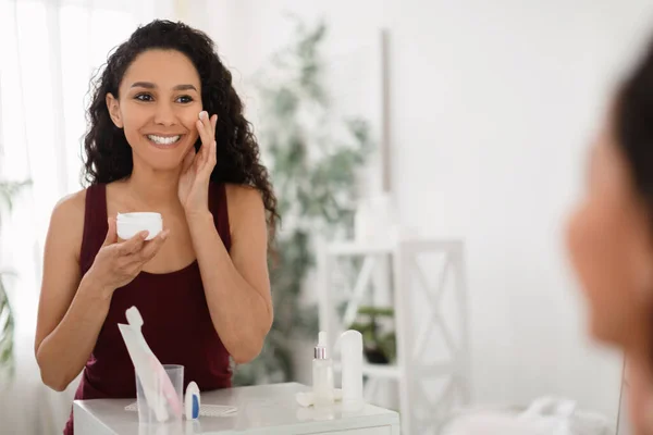 Mulher bonita aplicando creme facial perto do espelho em casa, espaço de cópia. Tratamento de spa, conceito de bem-estar — Fotografia de Stock