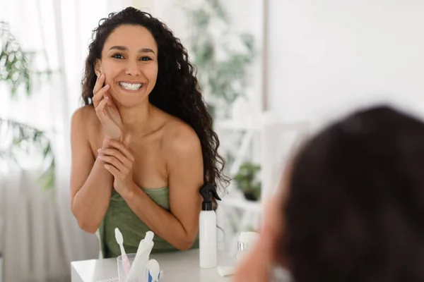 Gyönyörű boldog fiatal hölgy megérinti arcát tökéletes sima bőr közelében tükör a fürdőszobában, szabad hely — Stock Fotó