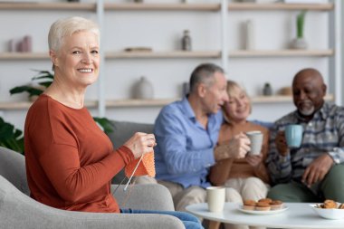 Neşeli kıdemli bayan örgü örüyor, çok ırklı arkadaşlarının yanında oturuyor.