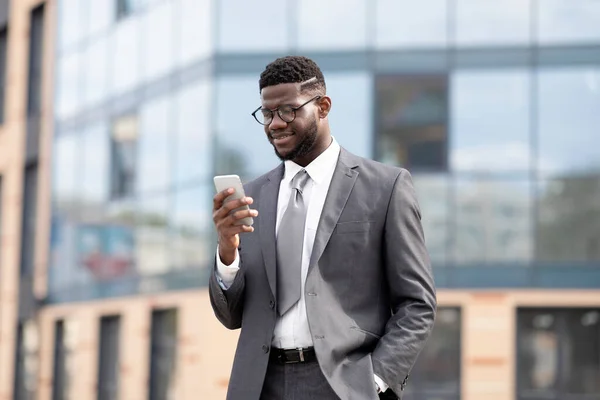 Administrador de TI negro exitoso en traje formal caminando al lado del edificio de oficinas, usando teléfono inteligente y sonriendo, espacio para copiar — Foto de Stock