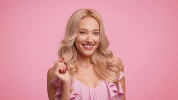 Blondýny žena hrát s kudrnaté vlasy usmívající se přes růžové pozadí — Stock video