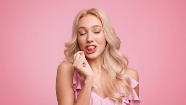 Блондинка Показ и поедание желе в форме сердца конфеты, розовый фон — стоковое видео