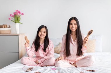 Mutlu genç Japon kadın ve pijamalı genç kız saç fırçaları ile yatakta oturup, yatak odasında eğleniyorlar.