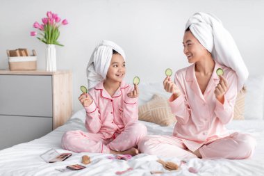 Tatmin olmuş bin yıllık Japon kadın ve genç kız pijama ve havluyla yatakta kozmetik ürünleriyle oturuyor, salatalık dilimleri tutuyor.