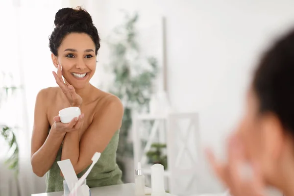 Hermosa mujer milenaria aplicando crema hidratante para la cara, de pie frente al espejo después de la ducha en casa, espacio para copiar — Foto de Stock