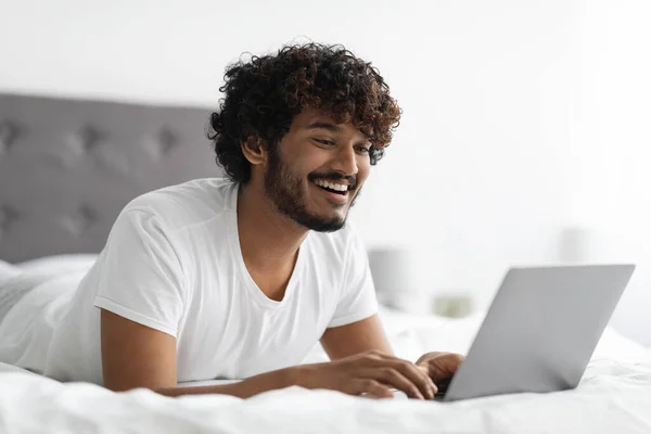 Fröhlicher indischer Mann tippt auf Laptop, arbeitet im Bett — Stockfoto