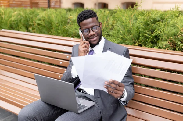 Εκτός γραφείου. African American επιχειρηματίας multitasking, μιλώντας σε smartphone και ανάγνωση εγγράφων, κάθεται στον πάγκο — Φωτογραφία Αρχείου
