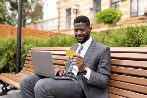 Homme d'affaires noir joyeux utilisant la carte de crédit et l'ordinateur portable, faire des achats en ligne tout en étant assis sur le banc — Photo