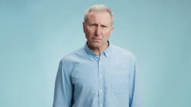 Öfkeli huysuz yaşlı adam kameraya yumruk atıyor, öfkeli ve öfkeli hissediyor, mavi stüdyo arka planı, yavaş çekim.