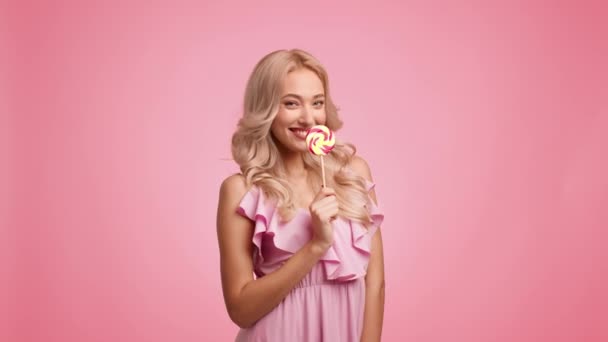 Блондинка леді їсть льодяник маючи задоволення позує грайливо, рожевий фон — стокове відео
