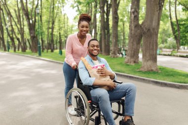 Tekerlekli sandalyedeki genç ve mutlu bir zenci, elinde bir çanta dolusu ürünle kız arkadaşıyla dışarıda alışverişe çıkıyor.