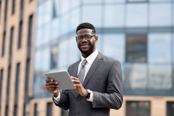 Θετικός Αφροαμερικανός επιχειρηματίας που χρησιμοποιεί ψηφιακή ταμπλέτα κατά τη διάρκεια του διαλείμματος γεύματος, περπατώντας κοντά στο κέντρο γραφείων σε εξωτερικούς χώρους — Φωτογραφία Αρχείου