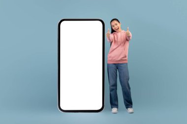 Neşeli Asyalı kadın büyük akıllı telefonuyla poz veriyor, başparmağını kaldırıyor.