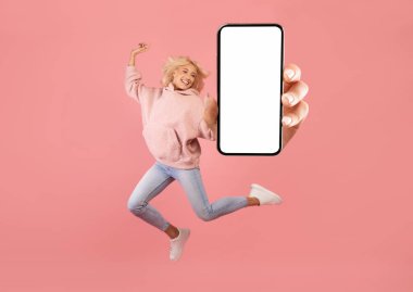 Pembe arka plan, kolaj, kopyalama alanının üzerinden atlarken boş cep telefonu ekranı gösteren aşırı neşeli bir kadın.