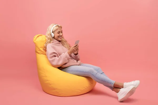 Mladá žena ve sluchátkách relaxující na fazolovém křesle, poslouchající hudbu nebo sledující film na mobilu — Stock fotografie