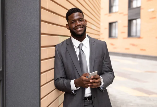 Glad afrikansk amerikansk entreprenör i kostym med smartphone, står nära kontorsbyggnad och ler mot kameran — Stockfoto
