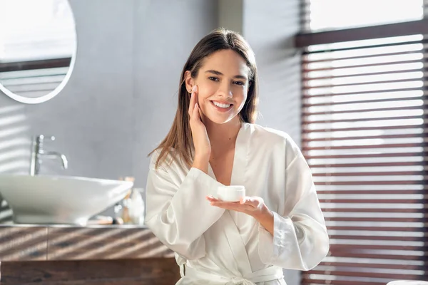 Concepto de Cosmética. Joven hermosa dama aplicando crema hidratante y sonriendo a la cámara —  Fotos de Stock