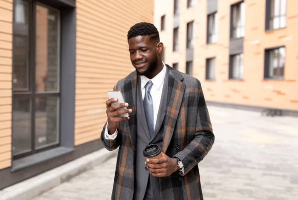 Glad svart affärsman sms:ar på mobilen och dricker hämtkaffe, går utanför kontorsbyggnaden — Stockfoto