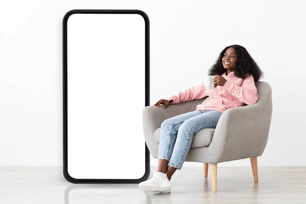 Lovely black woman sitting in armchair with coffee, offering space for your online ad, mobile app or website, mockup — Stock Photo, Image