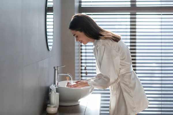 Mooie jonge vrouw dragen witte zijde mantel wassen gezicht in de badkamer — Stockfoto