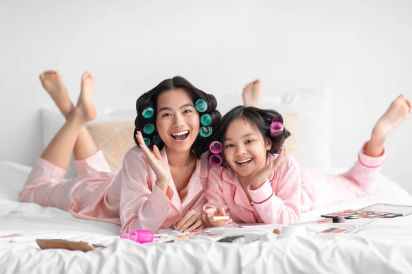 Felice felice millenaria signora giapponese e teen girl in pigiama e bigodini sdraiati sul letto con cosmetici, buon divertimento — Foto Stock