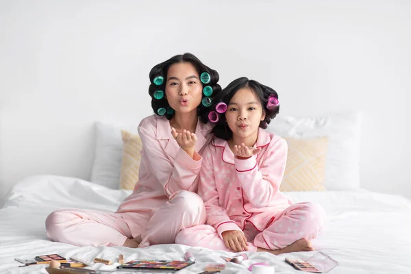 Lieto millenario signora giapponese e teen girl in pigiama e bigodini con cosmetici sul letto divertirsi e fare aria bacio — Foto Stock
