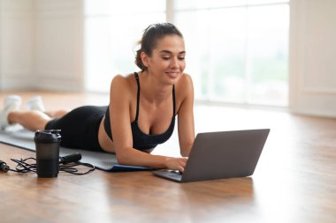 Gülümseyen genç bir kadın yoga minderinde dizüstü bilgisayar kullanarak yatıyor.