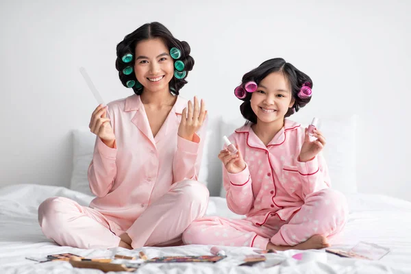 Sorridente millenario giapponese femmina e adolescente ragazza in pigiama e bigodini fare manicure sul letto in camera da letto — Foto Stock