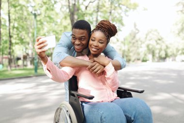 Tekerlekli sandalyedeki sevgilisine sarılan, parkta cep telefonuyla selfie çeken mutlu genç bir zenci.