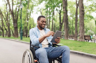 Tekerlekli sandalyedeki mutlu genç adam tablet bilgisayar kullanıyor, internette geziniyor ya da Spring Park 'ta film seyrediyor.