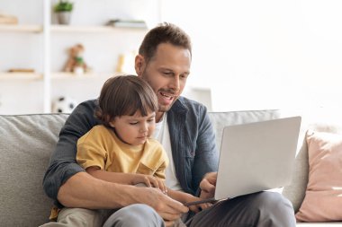 Aile işi konsepti. Evde dizüstü bilgisayarda çalışan genç baba iş adamı oğluna belgelerini gösteriyor ve açıklama yapıyor.