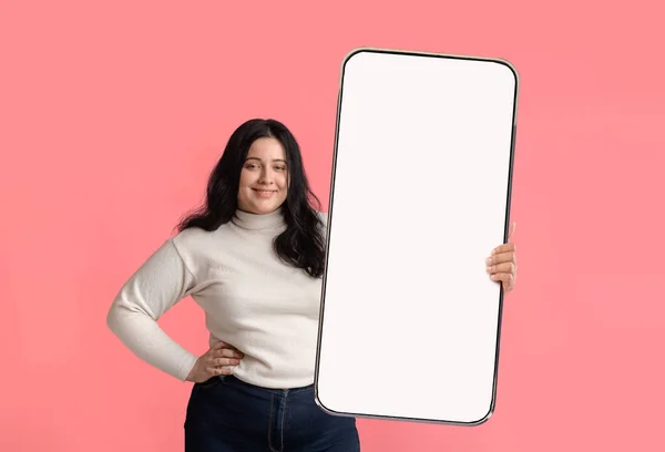 Усміхнена надмірною жінкою демонструє великий смартфон з порожнім білим екраном на камеру — стокове фото