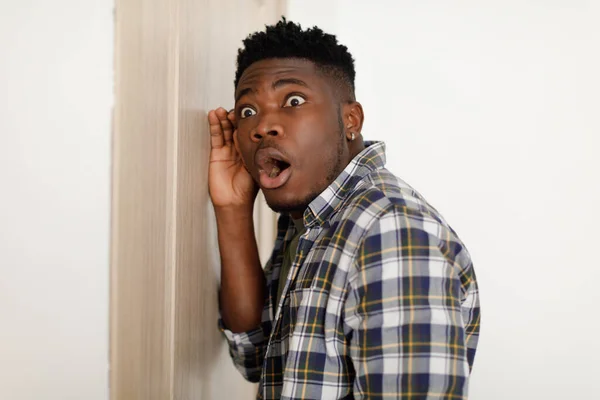 Zszokowany Afroamerykanin podsłuchujący przez zamknięte drzwi stojące w środku — Zdjęcie stockowe